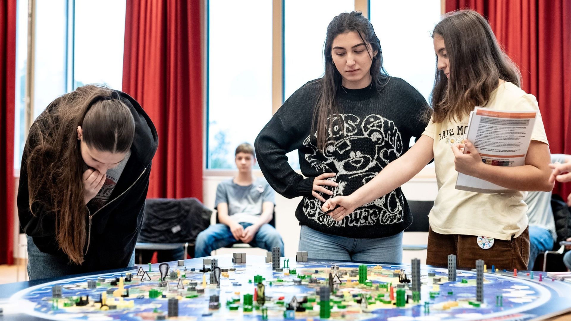 Schülerinnen und Schüler beugen sich über ein großes Weltklimaspiel-Spielbrett und diskutieren Spielzüge.