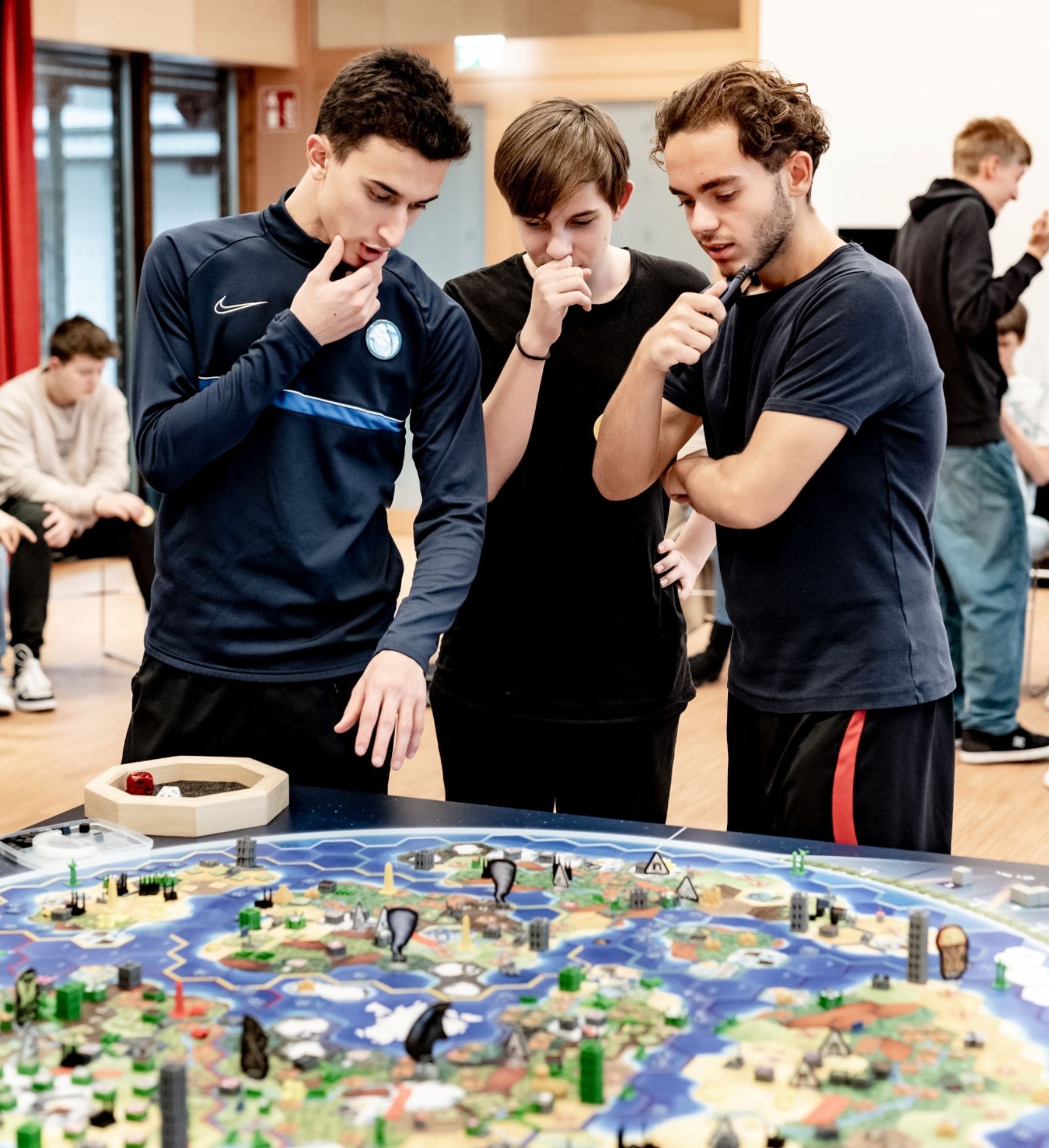 Drei Lehrlinge stehen &uuml;ber einem gro&szlig;en Weltklimaspiel-Brett und diskutieren gemeinsam eine Spielentscheidung.